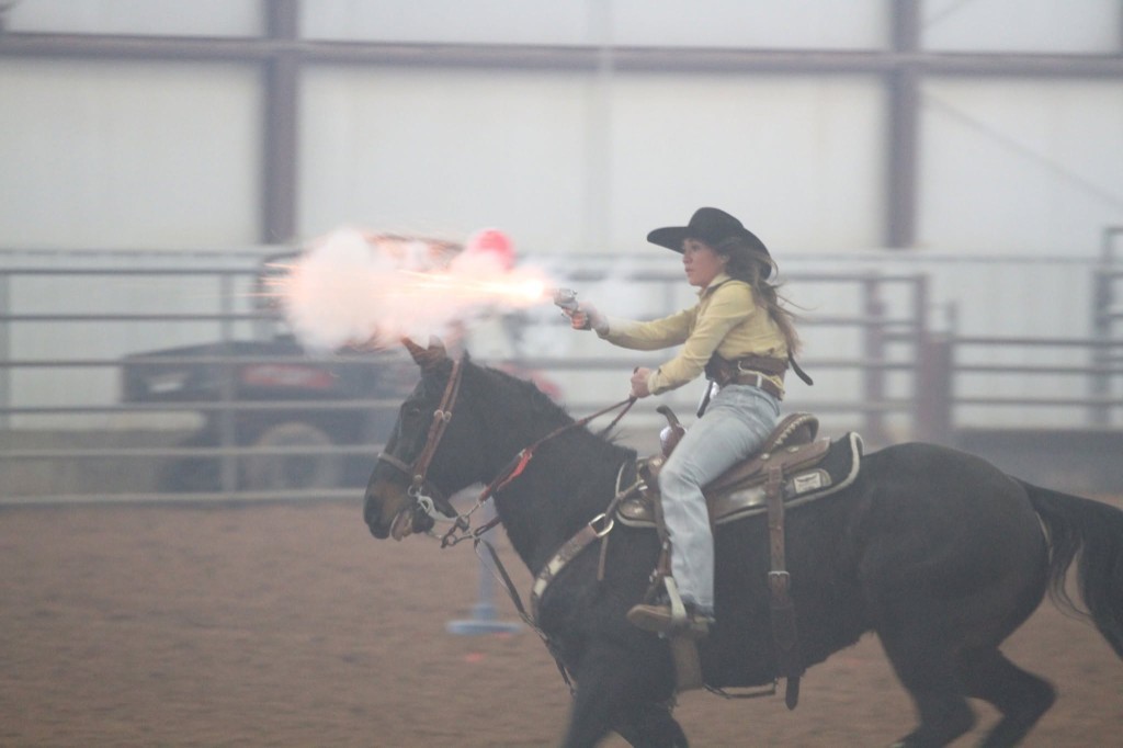 Mounted shooting rider in western attire inside the indoor arena.